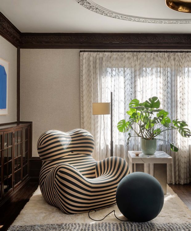 A richly detailed sitting room featuring a sculptural black-and-cream striped lounge chair, a dark spherical ottoman, a glass-front dark timber cabinet, a potted monstera, and sheer patterned curtains over a large window. The room has dark carved timber wall and ceiling trim, textured neutral walls, an ornate ceiling medallion, and a soft layered rug, creating a theatrical and refined interior.