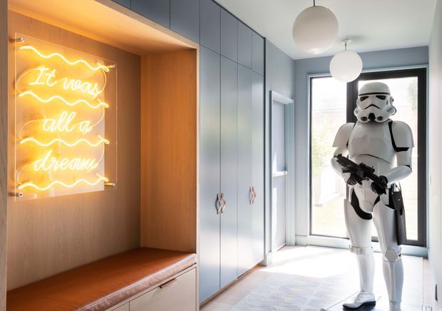 Bright, contemporary hallway or entry area with full-height pale blue-grey built-in cabinetry, warm timber-lined alcove seating, and a glowing neon sign. Large windows and spherical pendant lights add openness, while a Star Wars stormtrooper figure stands in the foreground.
