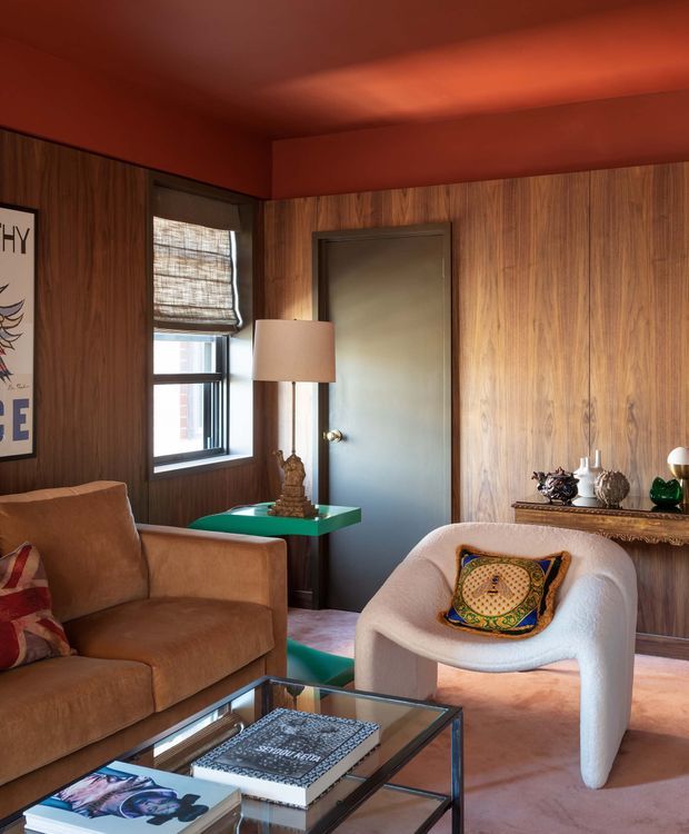 A richly coloured, eclectic living room with walnut-toned wood wall panelling, a deep rust-red ceiling and cornice, a dark painted internal door, and a small window with woven Roman blind. The room contains a caramel leather sofa, sculptural cream boucle armchair, green side table, glass-and-metal coffee table, decorative objects, and colourful cushions. Warm, layered lighting and saturated colours create a curated, intimate atmosphere.