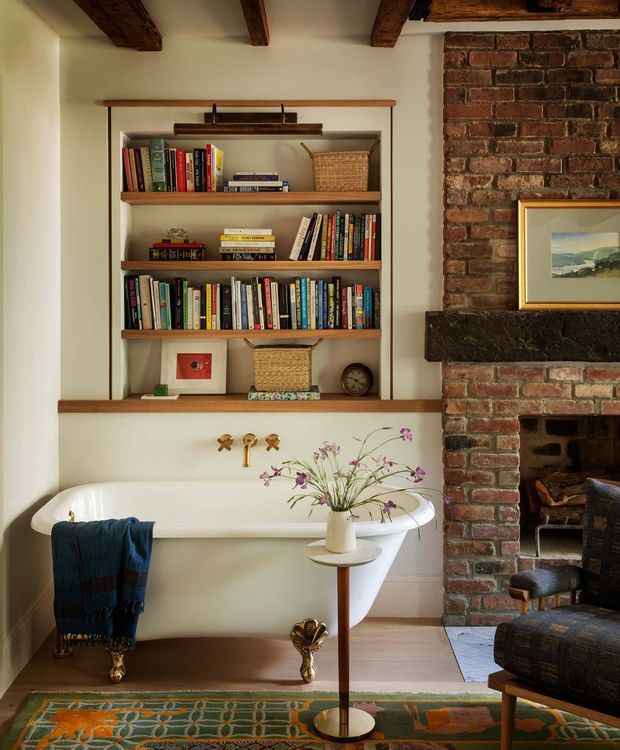 A warm, characterful bathroom featuring a freestanding white bathtub beneath built-in open timber shelving filled with books and baskets. The room includes exposed dark timber ceiling beams, pale plaster walls, a substantial exposed brick fireplace with dark stone mantel, brass wall-mounted bath fittings, timber flooring, and eclectic furnishings and accessories.