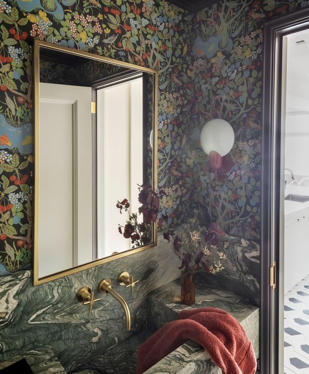 A richly styled powder room with dark botanical wallpaper, a dramatic green veined stone vanity backsplash and benchtop, wall-mounted brass tapware, and a large brass-framed mirror. A sculptural globe wall light and dried floral arrangement add decorative detail. The open doorway reveals part of an adjacent bright room with patterned flooring.