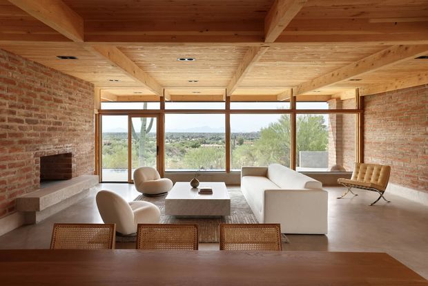 A spacious, warm-toned living room with exposed timber ceiling beams and timber-lined ceiling, exposed red-brick walls, and expansive timber-framed glazing overlooking a landscaped setting. A minimalist white sofa and armchairs surround a low stone or concrete coffee table, with a built-in brick fireplace along the left wall and a timber dining table in the foreground. The room has a refined mid-century-inspired architectural character with strong indoor-outdoor visual connection.