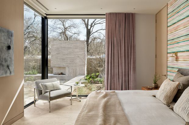 A refined contemporary bedroom with floor-to-ceiling corner glazing overlooking a landscaped courtyard and concrete structure. The room features a low-profile upholstered bed with layered neutral bedding, a sculptural lounge chair, pale built-in timber cabinetry, full-height dusty rose curtains, and a colourful horizontal striped feature wall. The palette is soft and warm with abundant natural light and a strong connection to the outdoors.