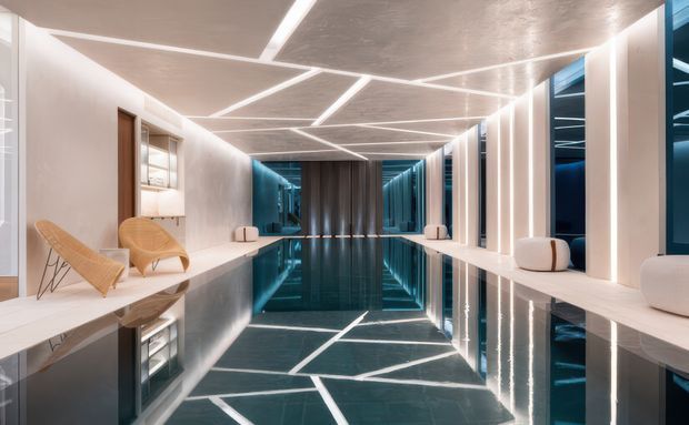 A dramatic, highly polished contemporary interior corridor or gallery with a reflective dark floor, white walls, and a textured ceiling featuring angular geometric linear lighting. Full-height glazing or mirrored blue-toned panels line the sides, while sculptural pale seating and warm timber lounge chairs add contrast. The symmetrical composition creates a luxurious, futuristic atmosphere.