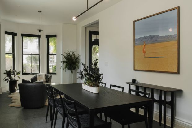 A refined open-plan dining and living area with a large dark rectangular dining table surrounded by black dining chairs, a slim black console, and black lounge seating. White walls, dark-framed windows, indoor plants, and a linear track light create a high-contrast contemporary interior. A large framed landscape artwork provides a warm earthy accent.