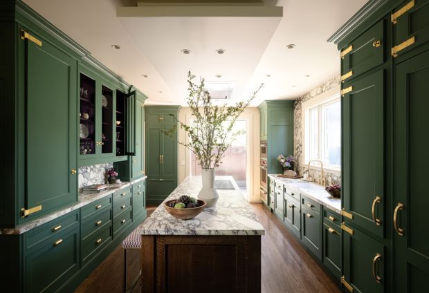 A bright, refined galley-style kitchen with deep green shaker-style cabinetry and extensive brass hardware. A central marble-topped island with a dark timber base anchors the room, while glass-front display cabinets, integrated storage, timber flooring, white walls, and large windows create a warm, classic interior. A skylight or roof window and recessed ceiling lights provide additional daylight and illumination.