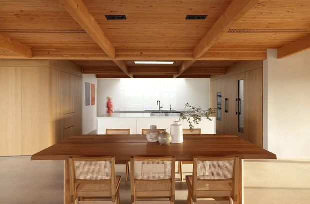Warm, open-plan dining and kitchen interior with a long dark timber dining table and three woven-cane dining chairs in the foreground. The kitchen beyond features a white island with integrated seating, pale timber cabinetry, dark benchtops, and stainless-steel appliances. Exposed timber ceiling beams and timber lining create a strong post-and-beam character, while broad glazing brings in soft natural light.