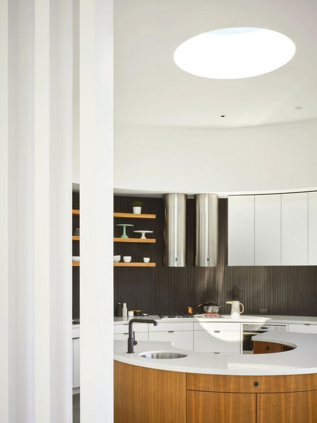 A bright, contemporary kitchen viewed through tall white structural columns. The space features a large curved island with white countertop and integrated sink, warm wood cabinetry, white wall cabinetry, dark vertically textured backsplash, open display shelving, stainless-steel cylindrical rangehoods, and a large oval ceiling light.