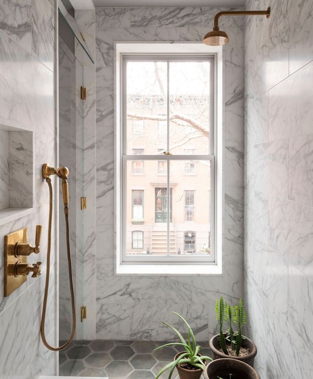 A compact, bright shower bathroom clad in veined white-and-grey marble-look wall tiles, with a frameless glass shower enclosure, brass shower fittings and rain shower, a tall sash window, and dark hexagonal floor tiles. Potted plants sit near the window and add a natural accent.
