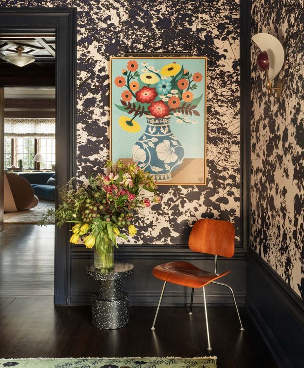 A richly styled interior vignette in a dark, highly decorative hallway or entry area. Black wall panelling and trim frame a dramatic cream-and-black abstract patterned wall covering. A colourful floral artwork hangs above a speckled black pedestal supporting a large flower arrangement. A vintage-style orange-brown upholstered chair with slim metal legs sits against the wall, with a sculptural wall sconce above. Through the doorway, a brighter adjoining living or dining space is visible. The room has dark timber flooring and an ornate, eclectic, high-contrast atmosphere.