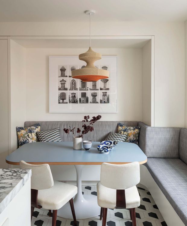 A compact, thoughtfully styled dining and lounge nook with a built-in grey upholstered banquette wrapping around a pale blue rounded dining table. Two cream upholstered dining chairs sit in the foreground, with patterned cushions, a sculptural woven pendant light, framed architectural artwork, and a bold geometric tiled floor. The space has a warm mid-century-inspired character with contemporary detailing.