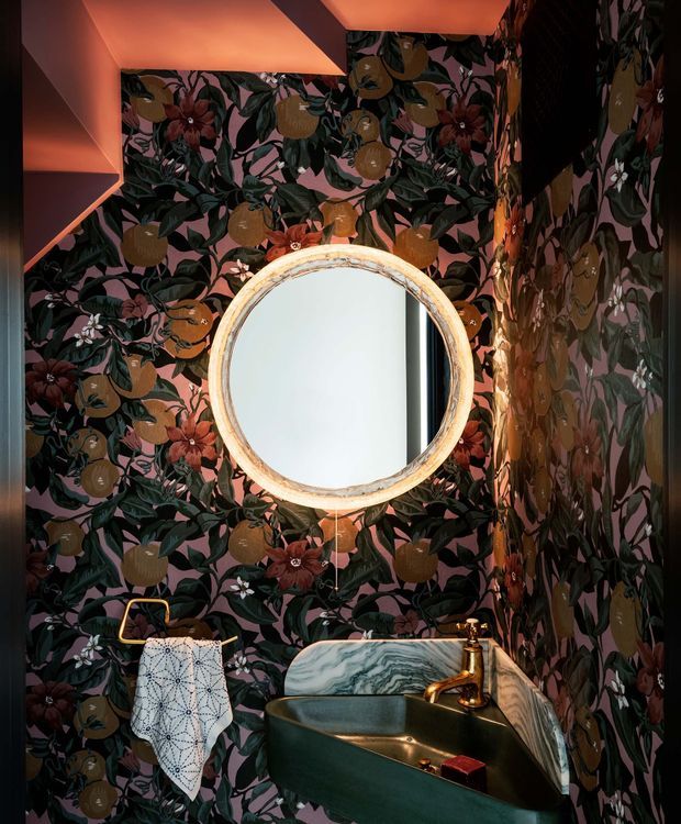 A compact, dramatically styled bathroom with dark botanical wallpaper featuring citrus fruit, leaves, and flowers across the walls. A round ornate framed mirror sits above a sculptural dark green basin with a marbled stone backsplash and polished brass tapware. Warm orange-pink concealed lighting illuminates the ceiling and upper walls, creating a rich, theatrical atmosphere. A patterned hand towel hangs from a brass rail beside the basin.