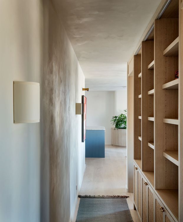 A narrow, softly lit residential hallway with a textured plaster wall, cylindrical wall sconces, pale timber flooring, and a full-height built-in timber shelving and cabinetry wall on the right. The corridor opens toward a brighter living area with a blue island or cabinet, a large potted plant, and minimal contemporary furnishings. The palette is warm and neutral with natural wood, off-white surfaces, and muted blue.