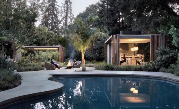A landscaped backyard centred on a curving swimming pool, with a paved pool surround, tropical and mature planting, a small outdoor lounge and fire feature, and a compact modern pavilion with timber-look vertical screening and an illuminated kitchenette or bar opening onto the pool area.