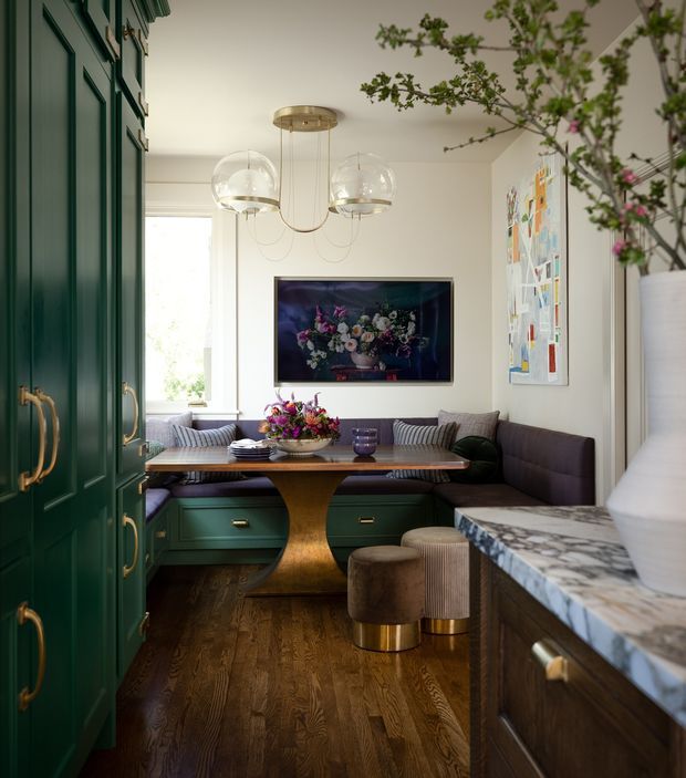 A refined, richly layered dining nook with a built-in banquette, dark green paneled cabinetry, and a central wooden dining table. The room features dark timber flooring, a marble-patterned stone benchtop in the foreground, brass hardware, sculptural glass pendant lighting, framed wall art, and a television. A large indoor branch arrangement adds an organic accent. The overall design is colourful, tailored, and contemporary with classic detailing.