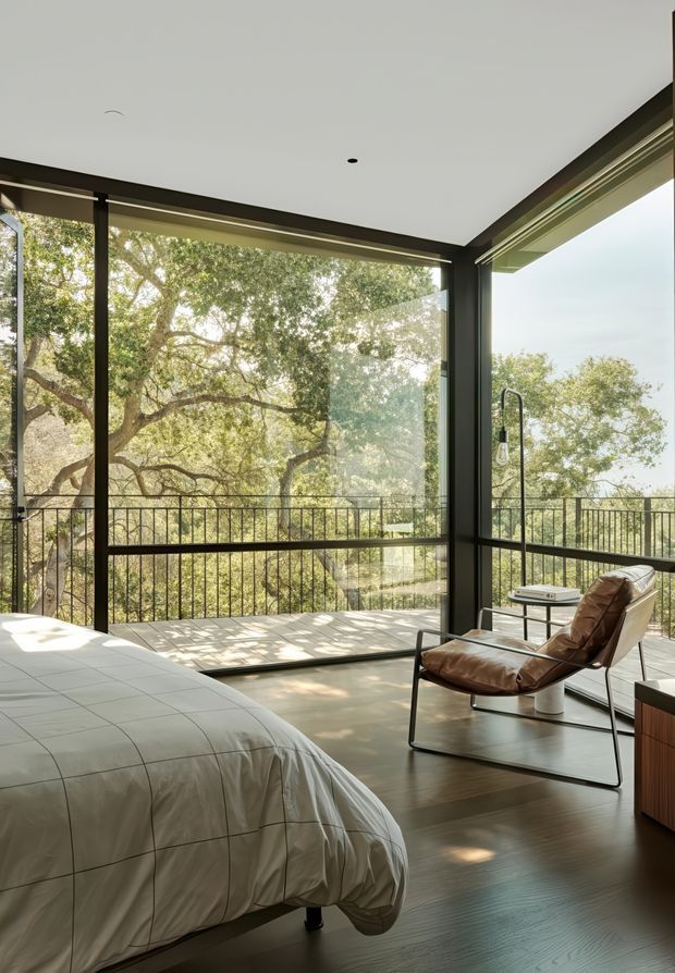 A contemporary bedroom with a large bed in the foreground, dark timber flooring, and floor-to-ceiling corner glazing opening onto a balcony. A leather sling lounge chair and small side table sit beside the windows, with abundant leafy trees visible outside. The room has a restrained palette of white, dark timber, black framing, and warm brown leather, creating a calm indoor-outdoor atmosphere.