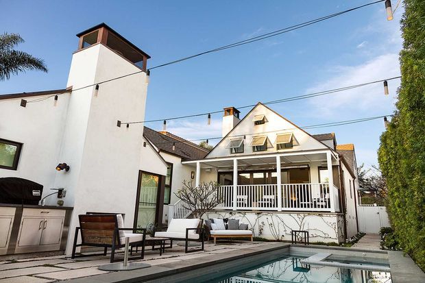 Exterior view of a multi-level residential home with white rendered walls, dark trim, steep gabled roof forms, dormer windows, and a covered veranda with white railings. A long swimming pool, paved terrace, outdoor kitchen, seating, festoon lighting, and dense hedging define the landscaped backyard entertaining area.