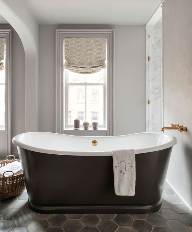 A refined bathroom centered on a freestanding black-and-white bathtub beneath a tall sash window. The room features pale grey walls with decorative layered window mouldings, a marble-look tiled wall and brass wall-mounted bath tapware. Dark hexagonal floor tiles, a woven basket and a patterned Roman blind add texture, while natural daylight creates a calm, elegant atmosphere.
