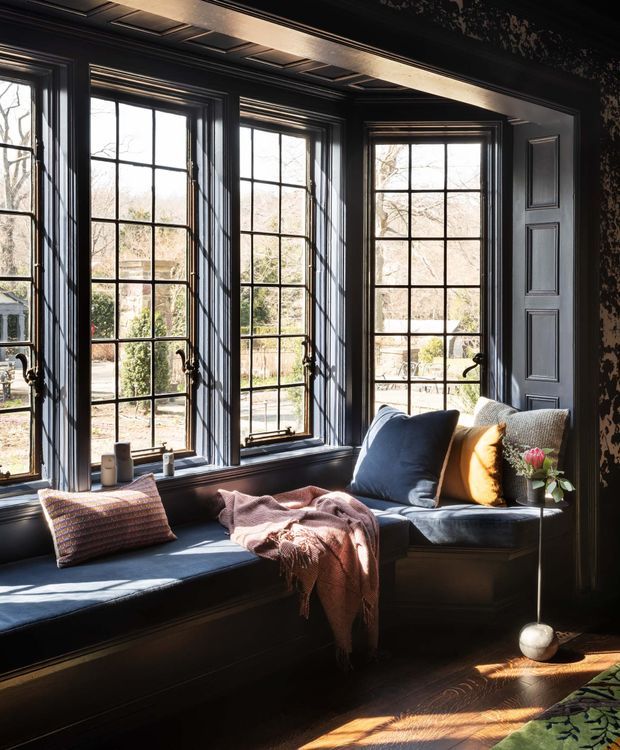 A refined window-seat nook beneath a row of tall, divided-light windows. The room features deep charcoal or black painted wall panelling, matching moulded window and door trim, a dark built-in bench seat, layered cushions, and a draped pink throw. Strong natural sunlight creates a warm, high-contrast atmosphere, while patterned wallpaper is visible at the upper right.