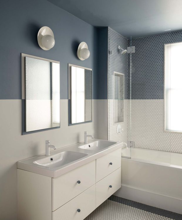 A bright, compact bathroom with a double white vanity, two integrated basins, wall-mounted faucets, and matching rectangular mirrors. The room features a white bathtub enclosed by clear glass, hexagonal mosaic tiles across the shower and bath walls and floor, blue-grey and white two-tone walls, and rounded wall sconces above the mirrors.