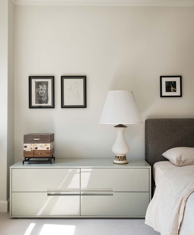 A softly lit bedroom vignette featuring a low white built-in or freestanding dresser with sleek handleless-style drawers, positioned beside a bed with a textured dark grey upholstered headboard. A sculptural white table lamp, framed monochrome artwork, and a small decorative storage box sit against a warm off-white wall. The room has a clean, restrained, contemporary feel with sunlight casting shadows across the cabinetry and pale floor.