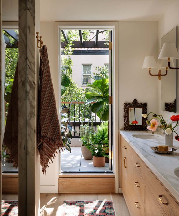 A warmly lit bathroom with a long light-wood vanity, polished white marble countertop, integrated basins, brass-toned hardware and fixtures, and a decorative dark-framed mirror. A tall glazed door opens onto a leafy balcony or terrace, bringing in abundant greenery and sunlight. Marble wall cladding and a striped hanging textile add texture, while a patterned rug and floral arrangements provide colour.
