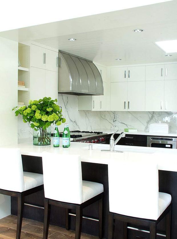 Bright, refined contemporary kitchen with a large white island and integrated seating for three. White cabinetry extends to the ceiling, with a dramatic curved stainless-steel range hood, marble-look backsplash, dark lower cabinetry, and recessed ceiling lighting. Dark timber bar stools and a vase of green flowers provide contrast against the predominantly white fit-out.
