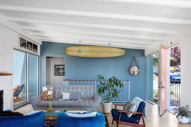 Bright coastal living room with white painted timber ceiling beams and walls, a blue vertical-panel feature wall, expansive windows and an open glazed door to a balcony or exterior. The room is furnished with a grey sofa, vivid blue lounge chairs, a timber coffee table, indoor plants, a surfboard mounted on the feature wall, and a round wall mirror. The space has an airy, relaxed beach-house character with abundant daylight.