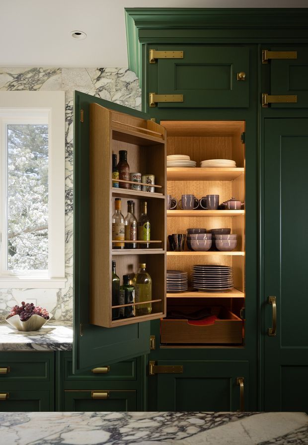 A close-up view of a richly detailed kitchen storage wall with deep green raised-panel cabinetry, brass hardware, and an open built-in pantry cabinet. The pantry has warm timber interiors, adjustable shelves, integrated lighting, plates, bowls, mugs, bottles, and a narrow spice and bottle rack on the inside of the door. A dramatic veined marble backsplash and matching countertop frame the cabinetry, with a white-trimmed window at left.