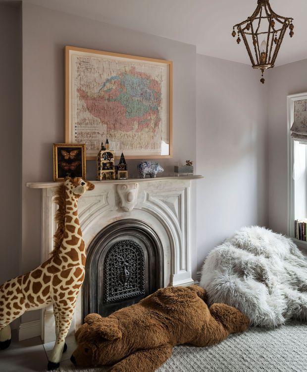 A richly styled traditional sitting room corner centered on an ornate white carved fireplace with a dark decorative fire screen. Above the mantel hangs a large framed vintage-style map, accompanied by small decorative objects and artwork. A sculptural antique-style pendant light hangs from the ceiling, while a window with a patterned Roman blind is visible at right. Plush animal-shaped furnishings, including a large giraffe and bear, and a shaggy white chair or throw create a playful, eclectic atmosphere. The room has muted mauve-grey walls, pale carpet, and refined historic detailing.