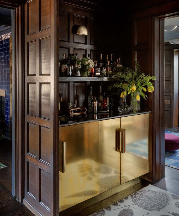A moody, richly detailed home bar recessed into dark timber wall panelling. The built-in bar features open bottle shelving, a black stone or dark reflective benchtop, and a brass-gold cabinet front with tall vertical handles. Floral arrangements, glassware, and bottles sit on the shelves and counter. A paneled timber door and surrounding dark wood trim reinforce the traditional, luxurious character, while glimpses of adjacent rooms appear through the openings.