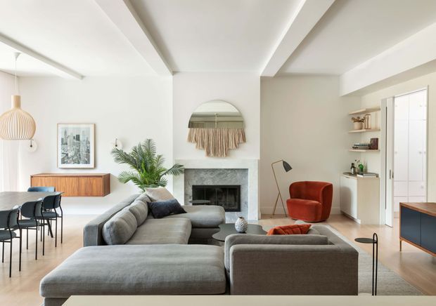 Bright, open-plan contemporary living and dining area with a large grey sectional sofa, timber dining table and black chairs, a red accent armchair, a marble or stone fireplace surround, built-in shelving, and pale neutral walls. Exposed ceiling beams and a central arch add architectural definition, while indoor plants, artwork, and woven wall decor provide warmth.