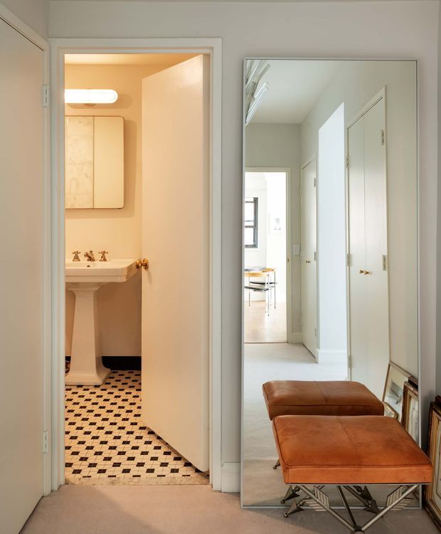 A bright residential hallway with a large full-height wall mirror reflecting the corridor and a distant dining area. An open door reveals a small traditional bathroom with a pedestal sink, wall-mounted mirror, warm lighting, and a black-and-white checkerboard tile floor. Two brown leather ottomans sit in front of the mirror, with framed artwork leaning along the wall.