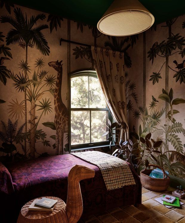 A richly decorated bedroom with dramatic botanical and animal-print wallpaper, a dark green ceiling, a tall arched-top window with patterned curtains, and abundant indoor plants. The bed is layered with a deep purple patterned coverlet and textured throws, accompanied by woven rattan furniture and warm ambient lighting from a large pendant fixture. The overall atmosphere is eclectic, moody, and richly patterned.