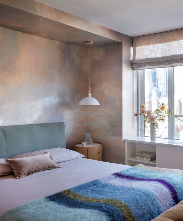 A stylish bedroom with a bed in the foreground, a softly upholstered blue-grey headboard, layered lavender bedding, and a colourful abstract throw. The room features a dramatic mottled metallic plaster feature wall and matching ceiling bulkhead, a small timber bedside table, a suspended white pendant light, and a large window with a Roman blind overlooking a dense city skyline. A built-in window seat or low shelving unit displays books and a vase of flowers.