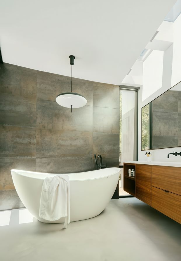 A bright contemporary bathroom featuring a freestanding white bathtub, large-format grey concrete-look wall tiles, a wall-mounted timber vanity with integrated white basin, black tapware, and a simple white pendant light. Tall glazing and a high angled ceiling bring in abundant natural light.