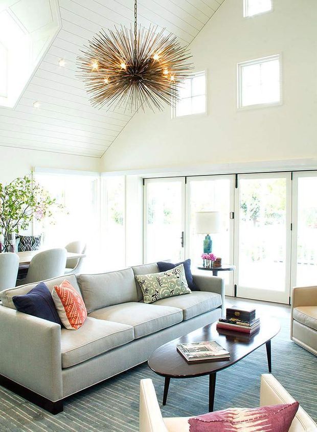 Bright, airy living room with a high vaulted white shiplap ceiling, extensive glazed folding or sliding doors, and a dramatic starburst pendant light. The room is furnished with a pale upholstered sectional sofa, patterned and coral accent cushions, a dark oval coffee table, armchairs, and a large blue-grey striped rug. A dining area and indoor plants are visible to the left.
