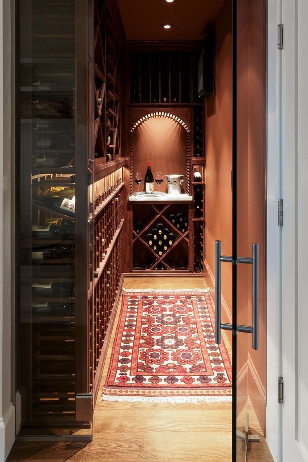 A compact, richly detailed wine cellar viewed through an open glass door. Dark timber wine racks and bottle storage line both sides, with a central tasting counter beneath an arched illuminated alcove. Warm terracotta walls, recessed ceiling lights, glass-fronted storage, and a patterned red oriental runner create an intimate, luxurious atmosphere.