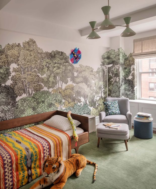 A playful, richly decorated bedroom with a low wooden daybed, colourful patterned bedding, a large botanical forest mural, and a green carpet. A grey upholstered armchair and footstool sit beside a window with a woven blind, while sculptural green pendant lights hang from the ceiling. A plush tiger toy and colourful hanging artwork add whimsical character.