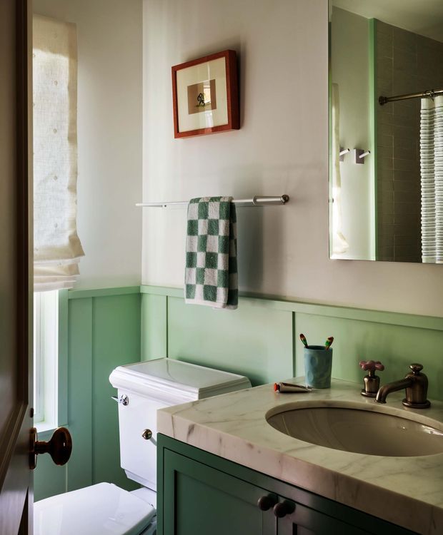 A compact, traditionally styled bathroom with pale green wainscoting and matching vanity cabinetry, a marble benchtop with an inset oval basin, bronze-toned tapware, white toilet, towel rail, framed artwork, and a window with a Roman blind. A mirror reflects the tiled shower area and another towel rail.