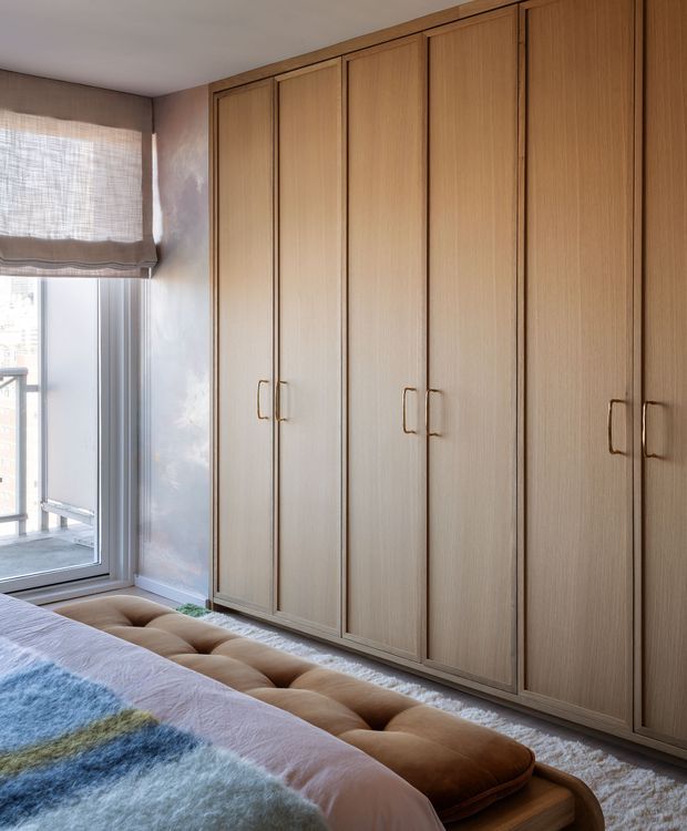 A softly lit bedroom dominated by a full-height wall of warm light-wood built-in wardrobes with vertical panelled doors and slim metal handles. A bed with plush layered bedding and a tufted brown upholstered bench occupies the foreground. A large glazed window with a textured Roman blind brings in daylight, and a pale textured carpet and muted neutral finishes create a calm, refined atmosphere.