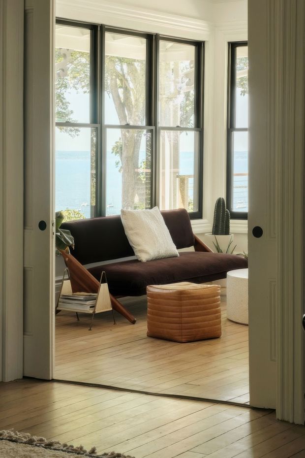 A bright, refined sitting room viewed through open double doors, featuring a dark brown mid-century-style sofa with a cream cushion, a rounded tan upholstered ottoman, a small sculptural side table, and potted cacti. Tall black-framed windows overlook blue water and trees, while pale timber flooring, white walls, and detailed ceiling mouldings create an airy coastal setting.