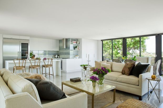 A bright open-plan living and dining area connected to a contemporary white kitchen. The room features large black-framed windows, cream sofas, a glass-topped coffee table with a timber-toned frame, light flooring, white cabinetry, and a blue-grey tiled splashback. Natural greenery is visible outside the glazing.