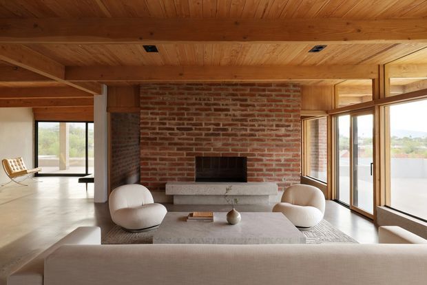 A warm, open-plan living room centered on a full-height exposed red-brick fireplace with a dark firebox and pale stone hearth. The room has extensive timber wall and ceiling lining, exposed timber structural beams, large timber-framed glazing and sliding doors, pale polished flooring, a low neutral sofa, rounded lounge chairs, and a simple coffee table. Outdoor views are visible through the glazing.