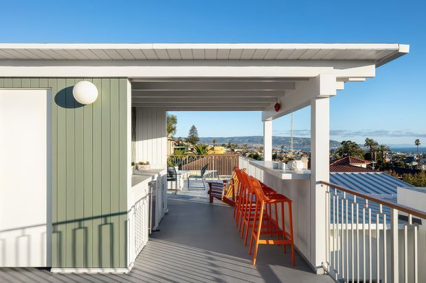 A bright rooftop terrace or balcony with a covered outdoor bar and a row of vivid orange bar stools. The space features pale grey decking, white structural posts and balustrades, green vertical cladding, exposed roof framing, and broad coastal or urban views beyond the railing.