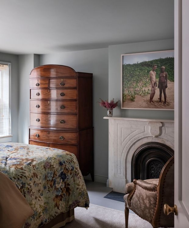 A refined traditional bedroom with a tall dark timber antique chest of drawers, a floral upholstered bed, a carved white fireplace surround with a dark arched insert, and a classic upholstered armchair. Pale grey walls, a light carpet, and framed artwork create a calm, historic interior atmosphere.