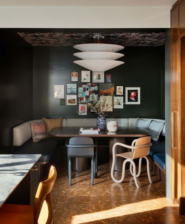 A richly styled dining nook with a built-in dark banquette wrapping around a wooden dining table, surrounded by sculptural chairs. The room features a dramatic black wall with a gallery arrangement of framed artwork, a layered white pendant light, timber cabinetry or panelling at the right, and warm patterned stone or timber flooring. Strong sunlight enters from the foreground, creating a warm, intimate atmosphere.