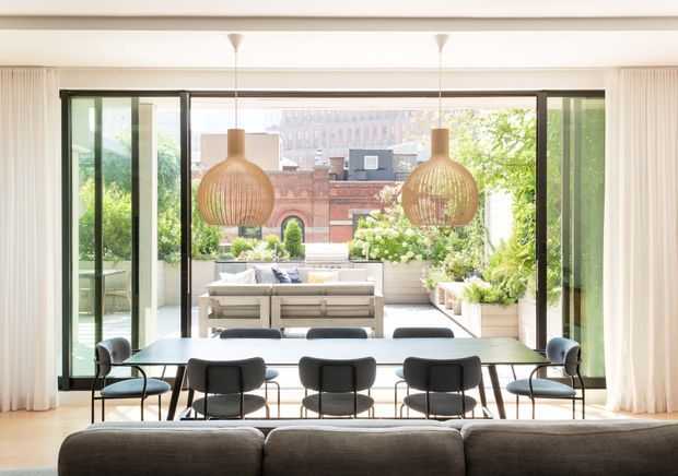 Bright, refined dining and living space opening onto a lush urban terrace. A long dining table with contemporary dark chairs sits beneath two woven pendant lights, while floor-to-ceiling glazed sliding doors and sheer curtains create a strong indoor–outdoor connection. A sofa is visible in the foreground, and the terrace includes built-in seating, planters, and views toward surrounding city buildings.