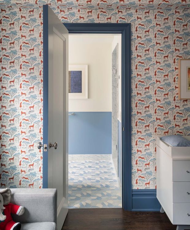 A whimsical, highly patterned bedroom or dressing area with animal-and-tree wallpaper in white, blue and red. A deep blue painted door frame and skirting contrast with dark timber flooring. Through the open door is a bright bathroom featuring pale blue lower walls, white upper walls, a patterned blue-and-white tile floor, and marble-look wall tiling. A white chest of drawers and upholstered grey seating are visible in the foreground.