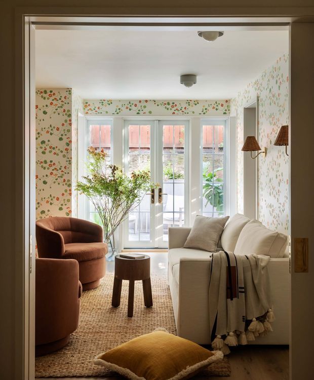 A bright, compact sitting room viewed through a wide doorway, with a cream sofa, rust velvet armchairs, a small round timber stool, woven natural-fibre rug, and mustard floor cushion. Floral patterned wallpaper covers the walls, while pale blue-grey French doors and tall windows open to a small exterior balcony or courtyard. Wall-mounted shaded sconces and simple flush ceiling lights complete the warm, traditional interior.